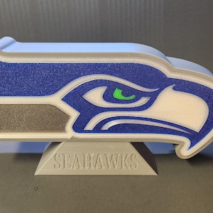 Könnte beinhalten: Ein 3D-gedrucktes Seahawks-Logo in Blau, Weiß, Grau und Grün. Das Logo ist ein stilisierter Falkenkopf, montiert auf einem grauen Sockel mit dem Wort "SEAHAWKS" in Großbuchstaben. Es ist ein Fanartikel.