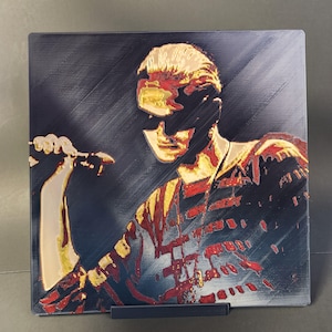May include: A square, textured print of a person holding a microphone, with a dark background. The person is wearing sunglasses and a shirt with red and gold accents. The print is displayed on a black stand.