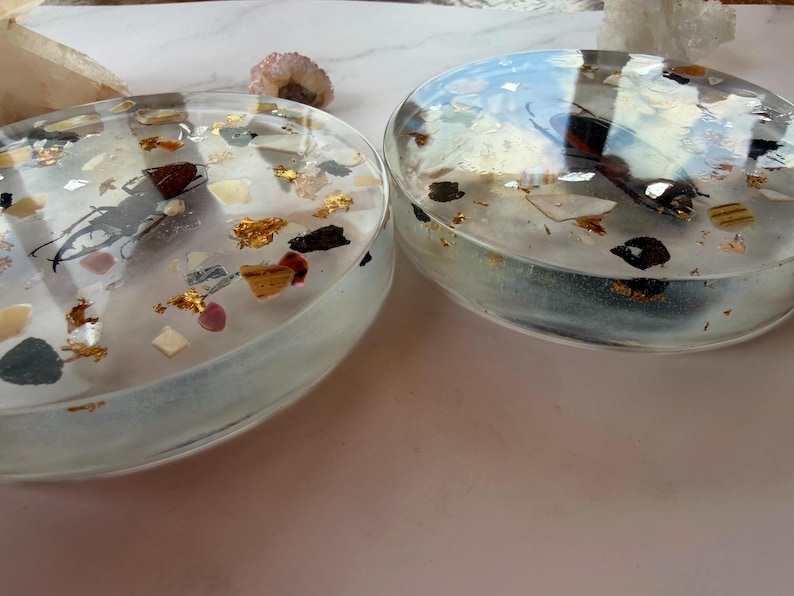 May include: Two clear, oval-shaped resin coasters. Each coaster contains gold leaf, small pieces of stone, and a dark insect. The coasters are on a light pink surface.