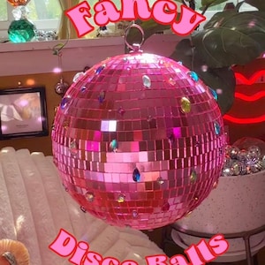 May include: A spherical disco ball covered in small pink mirrored tiles and accented with various colorful, faceted rhinestones. A metal ring is attached to the top for hanging. The text "Fancy" and "Disco Balls" is overlaid in pink letters.