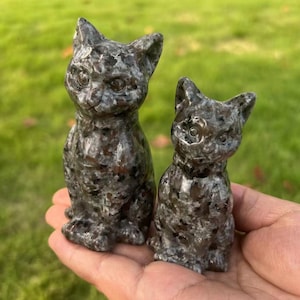 Puede incluir: Dos figuritas de gato de piedra pulida, con manchas negras y grises. El gato más grande está sentado erguido con las orejas levantadas. El gato más pequeño está sentado con la cabeza inclinada hacia un lado.
