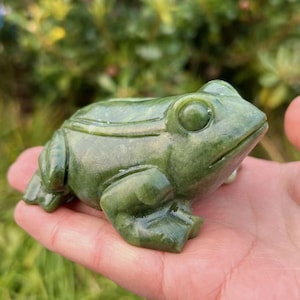 Könnte beinhalten: Eine grüne Jade-Froschfigur mit detaillierter Schnitzerei. Der Frosch sitzt auf einer Hand mit geschlossenen Augen.