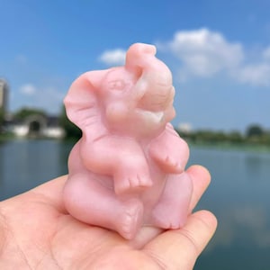 May include: A pink, hand-carved elephant figurine made of rose quartz. The elephant is sitting with its trunk raised and its legs tucked underneath its body.