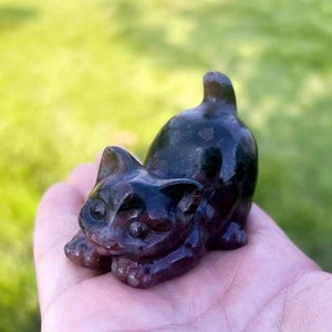 Puede incluir: Una pequeña figurita de gato negro y verde tallada en piedra pulida. El gato está acostado sobre su vientre con la cabeza girada hacia un lado.