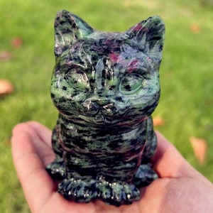 Puede incluir: Una figurita de gato verde y negro tallada en una piedra preciosa. El gato está sentado con las patas metidas debajo de él. La figurita mide aproximadamente 7,5 cm de alto.
