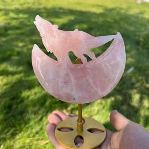 Puede incluir: Un cristal de cuarzo rosa tallado en forma de lobo aullando a la luna. El cristal está montado en un soporte de metal dorado.
