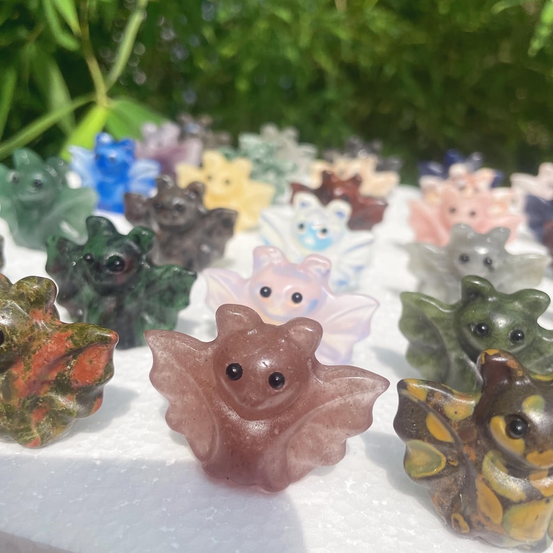May include: A collection of gemstone bat figurines in various colors, including green, brown, pink, and blue. Each bat has detailed wings and a small, round body. The bats are arranged closely together, creating a whimsical display.