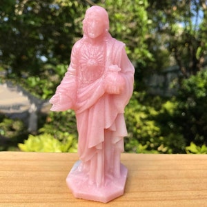 May include: A pink resin statue of Jesus Christ with a heart on his chest and his hands clasped in prayer.