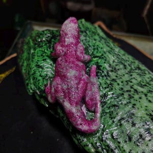 May include: A textured, magenta-colored lizard figurine with silver-toned highlights resting on a larger, irregularly shaped, vibrant green rock with black speckles. The lizard is positioned vertically, showing its back, head, and curled tail.