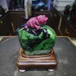 May include: A green and black gemstone carved into the shape of a frog sitting on a green and black rock. The frog is made of pink gemstone. The rock and frog are on a wooden base.