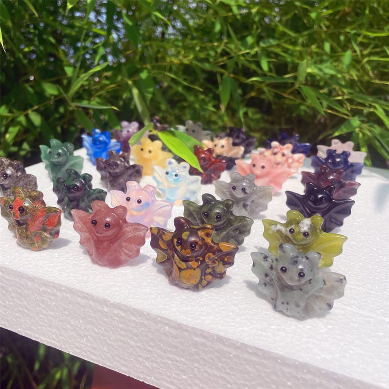 May include: A collection of small, carved bat figurines in various colors, including red, pink, green, and yellow. The bats are displayed on a white surface against a backdrop of green foliage. Each bat has detailed wings and facial features.