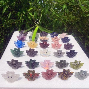 May include: A collection of small, carved crystal bat figurines in various colors, including blue, pink, green, and purple. The bats have outstretched wings and detailed features, displayed on a white surface.