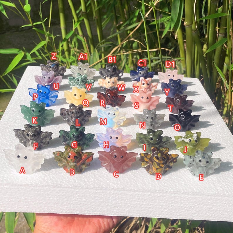 May include: A collection of small, carved bat figurines in various colors, including green, pink, and brown. Each bat has outstretched wings and is displayed on a white surface. The figurines are labeled with letters and numbers.