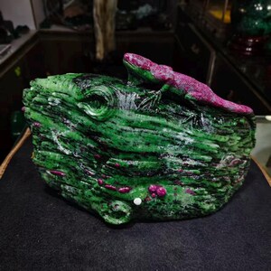 Könnte beinhalten: Eine geschnitzte Steinskulptur, die eine Eidechse mit einem leuchtend magentafarbenen Körper auf einer strukturierten, dunkelgrünen Felsbasis zeigt. Der grüne Stein weist natürliche schwarze Einschlüsse und magentafarbene Mineralablagerungen auf.