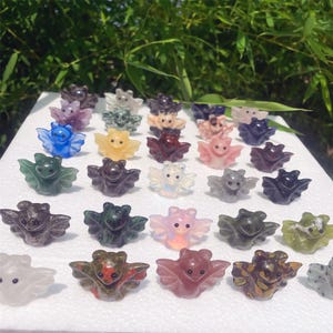 May include: A collection of small, carved bat figurines in various colors, including blue, green, yellow, and pink. The bats are made of polished stone and have outstretched wings. The figurines are arranged on a white surface.