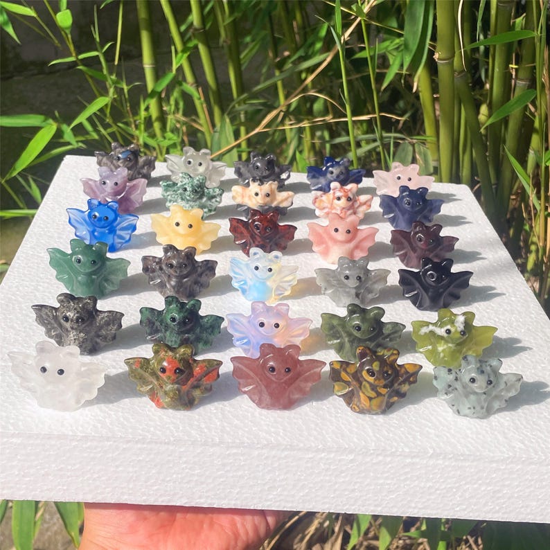 May include: A collection of small, carved bat figurines in various colors, including blue, green, pink, and brown. The bats are displayed on a white surface, with a natural green background.
