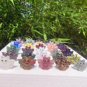 May include: A collection of small, carved crystal bat figurines in various colors, including green, pink, purple, and clear. The bats are arranged on a white surface against a backdrop of green foliage. Each bat has outstretched wings and a smiling face.