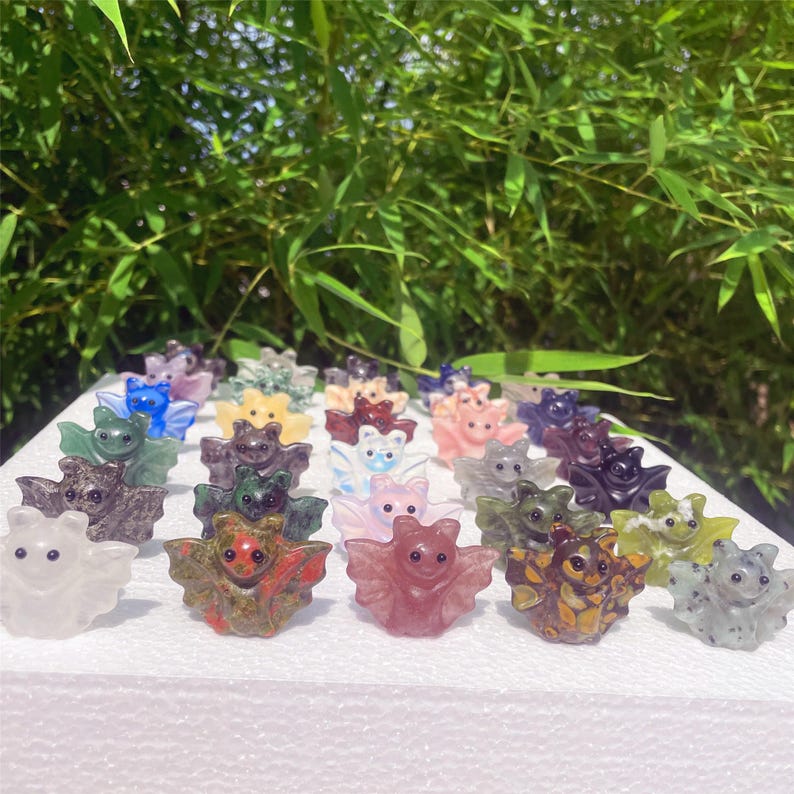 May include: A collection of small, carved crystal bat figurines in various colors, including green, pink, purple, and clear. The bats are arranged on a white surface against a backdrop of green foliage. Each bat has outstretched wings and a smiling face.