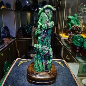 Puede incluir: Escultura tallada en piedra verde y roja de un dragón enrollado alrededor de un pilar. La escultura está montada sobre una base redonda de madera oscura. El dragón y el pilar tienen una superficie texturizada.