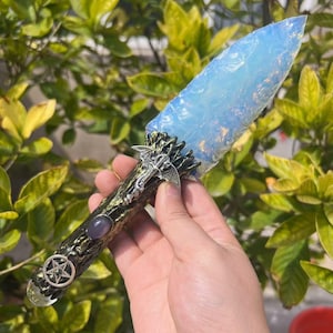 May include: A decorative dagger with a light blue, opal-like blade. The handle is dark green with silver dragon accents and a pentagram symbol. A purple stone is set into the handle near the blade.