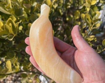 May include: A carved banana sculpture in shades of yellow and cream. The banana is held in a hand, with a blurred green leafy background. The sculpture has a smooth, polished appearance, with subtle variations in color.