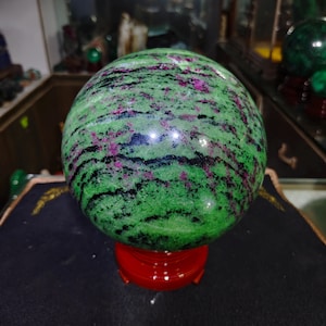 Può includere: Una pietra sferica lucida caratterizzata da una base verde vibrante con striature prominenti di inclusioni minerali color magenta intenso e nero. La sfera è montata su un supporto circolare rosso lucido, con una superficie liscia e riflettente.