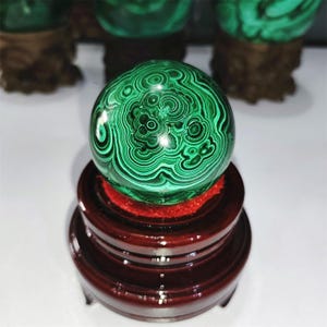 May include: A green malachite sphere with swirling patterns sits on a dark wood stand with a red felt lining.