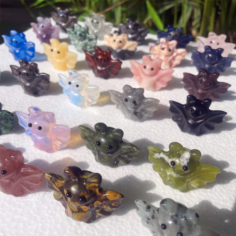 May include: A collection of small, carved gemstone bat figurines in various colors, including blue, pink, green, and black. The bats have outstretched wings and detailed features, perfect for a unique gift or decorative item.