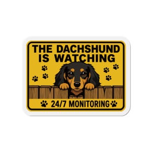 May include: A rectangular yellow sign featuring a black and tan dachshund peeking over a wooden fence. The text reads THE DACHSHUND IS WATCHING at the top and 24/7 MONITORING at the bottom, accented by black paw prints on the yellow background.