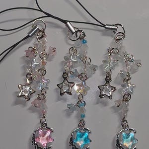 May include: Three phone charms featuring silver-tone metal hardware, dangling iridescent star-shaped beads, and small flower-shaped beads. Each charm ends with a silver crescent moon pendant containing a translucent pink, blue, or iridescent star.