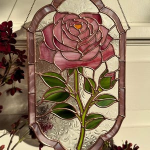 Op de afbeelding: Gebrandschilderd glaspaneel met een roze roos met groene bladeren en stengel. De roos heeft een oranje centrum. Het paneel heeft een decoratieve rand en is ontworpen om op te hangen. Het achtergrondglas heeft een bloemmotief.