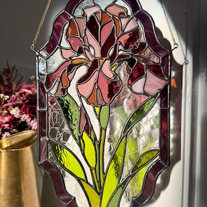 May include: Stained glass panel featuring an iris flower design. The panel has a dark purple border and a gold chain for hanging. The flower is depicted in shades of pink, red, and orange, with green leaves and a yellow stem. The glass has a textured appearance.