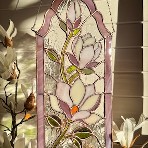 May include: A stained glass window hanging featuring a floral design with two blooming magnolia flowers and a bud. The piece is crafted with pale pink, white, and green glass segments held together by copper foil lines, framed in a pointed arch.
