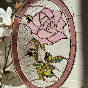 Peut inclure: Un panneau ovale en vitrail représentant une rose rose avec des feuilles et une tige vertes. La rose est le point central, sur un fond clair texturé. Le panneau est encadré dans un métal couleur cuivre.