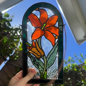 Puede incluir: Panel de vidrieras con un lirio naranja vibrante con hojas verdes y un capullo. La flor se encuentra sobre un fondo claro y texturizado dentro de un marco verde oscuro y negro. Una pieza de arte floral decorativa.