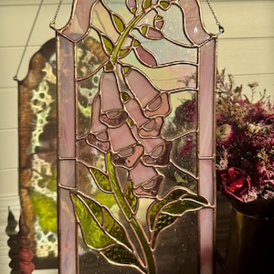 Könnte beinhalten: Ein Buntglasfenster mit einem Motiv aus rosa Fingerhutblüten und grünen Blättern, eingefasst in einen kupferfarbenen Metallrahmen. Das bogenförmige Stück hängt an einer dünnen Metallkette und zeigt durchscheinende Glasstrukturen in Rosa und Grün.