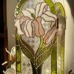 May include: Stained glass panel featuring a stylized lily flower in shades of white, pink, and green. The flower is set against a textured background, framed by a green border and copper-colored lines. The panel is suspended by a chain.