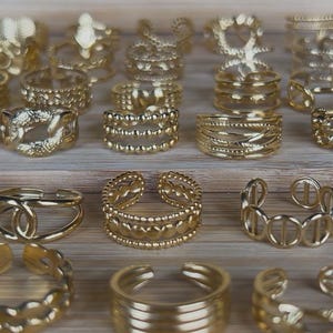 May include: A collection of gold-colored rings in various designs, including open-ended, band, and statement styles. The rings are displayed on a light-colored wooden surface, showcasing their intricate details and textures. The rings are made of a shiny, metallic material.