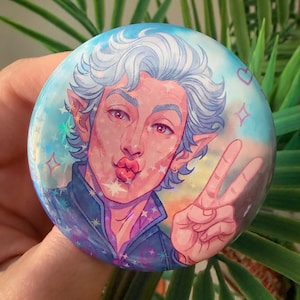 Könnte beinhalten: Ein runder Button mit einem Porträt einer Person mit weißen Haaren, roten Augen und spitzen Ohren. Die Person macht einen Kussmund und zeigt das Friedenszeichen. Der Button hat einen blau-rosa Hintergrund mit Sternen und Herzen.