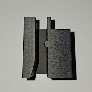 May include: A dark gray, textured metal object with a unique shape. It features a rectangular base with a vertical, flat piece and a curved indentation. The object has a matte finish and appears to be a component or part of a larger structure.