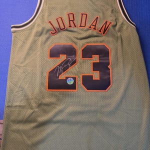 May include: Olive green basketball jersey with the name "JORDAN" in orange lettering above the number "23". The number is black with an orange outline. The jersey has a white and olive green trim.
