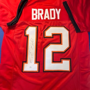 May include: Red football jersey with the name "BRADY" across the shoulders and the number "12" on the front and back. The jersey has a white and orange trim and a signature on the front.