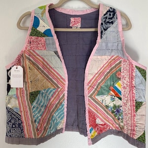 May include: A patchwork vest with a pink trim and a purple lining. The vest is made of various fabrics, including stripes, floral prints, and paisley. The vest is hanging on a wooden hanger.