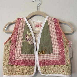 May include: A quilted vest with a patchwork design featuring floral and plaid patterns in shades of pink, green, and white. The vest has a white trim and a V-neck.