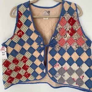 May include: A blue quilted vest with a patchwork design featuring red, white, and blue squares. The vest has a tie closure at the front.