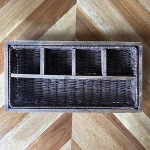May include: A rectangular, dark brown woven wicker basket with a wooden frame. The basket is divided into three compartments by vertical wooden dividers and a horizontal shelf. The basket sits on a wooden surface with a chevron pattern.