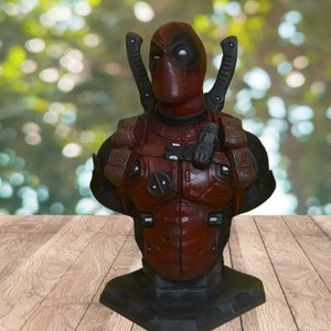 Op de afbeelding: Een rode en zwarte Deadpool-bustefiguur met een gedetailleerde sculptuur van zijn kostuum, inclusief zijn kenmerkende zwaarden en masker. De buste wordt gepresenteerd op een zwarte basis.