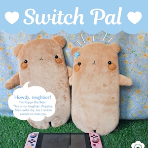 Puede incluir: Dos fundas Switch Pal con forma de oso de peluche marrón, con rasgos bordados y brazos pequeños. Una consola Nintendo Switch rosa descansa debajo. El texto dice "Switch Pal" y "Howdy, neighbor!" sobre un fondo azul claro.