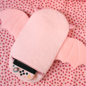 May include: A pink plush bat-shaped case for a Nintendo Switch console. The case has a zipper opening and a fuzzy texture.