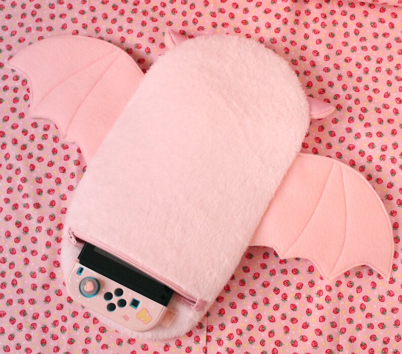 May include: A pink plush bat-shaped case for a Nintendo Switch console. The case has a zipper opening and a fuzzy texture.